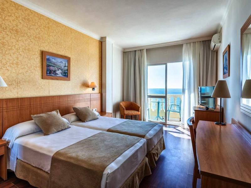 Helles Hotelzimmer mit zwei Betten, Holzmöbeln und Balkon mit Meerblick bei Tageslicht.