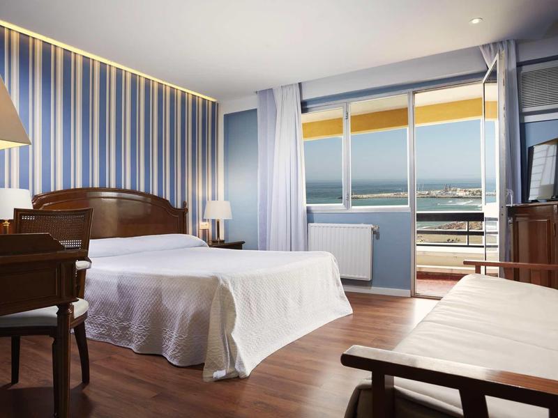 Helles Hotelzimmer mit Doppelbett, Holzmöbeln, Balkon und Meerblick bei Sonnenuntergang.