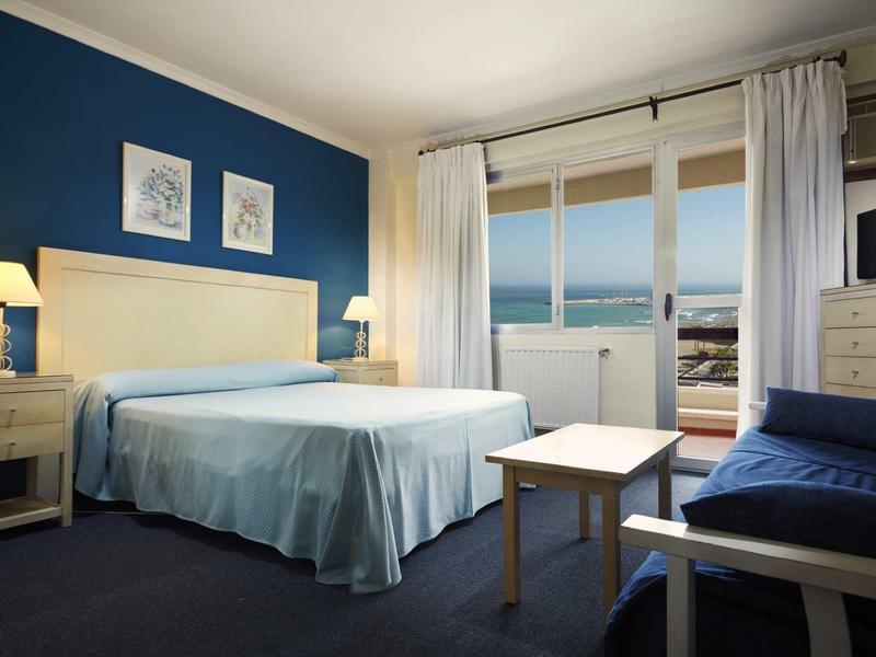 Schlafzimmer mit großem Bett, blauer Wand, weißen Vorhängen und Blick auf das Meer.