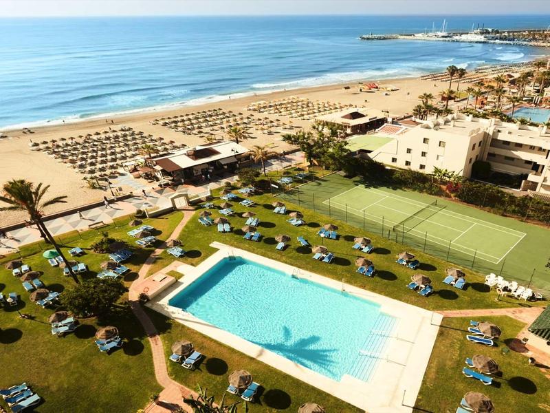 Blick auf Hotelanlage mit Schwimmbad, Liegestühlen, Palmen und Strand am blauen Meer.