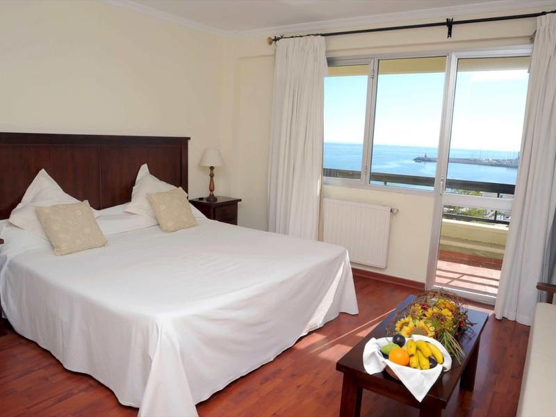 Hell eingerichtetes Schlafzimmer mit Doppelbett, Holzmöbeln und Blick auf das Meer durch Glasfenster.