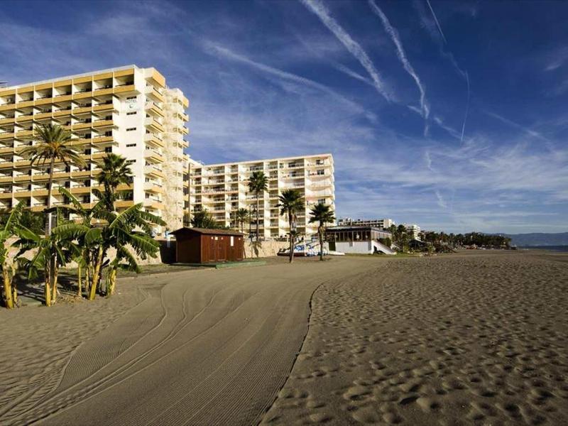 Großer Sandstrand mit Palmen vor mehrstöckigen weißen Hotelgebäuden unter blauem Himmel.