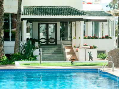 Hotelgarten mit klar blauem Pool, Treppe und Eingang zu einem weißen Gebäude mit grüner Dachterrasse.