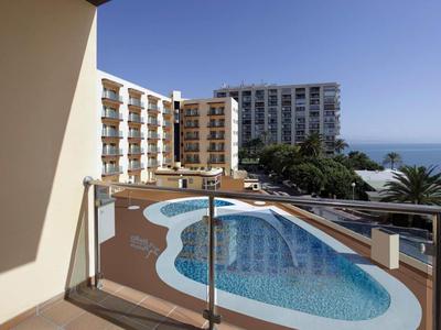 Blick vom Balkon auf geschwungenen Pool, Hotelgebäude und blauen Himmel mit Meer im Hintergrund.