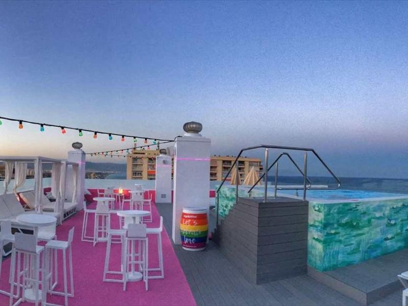 Dachterrasse mit Pool, Sonnenliegen, hohen Tischen, bunten Sitzkissen und Beleuchtung bei Dämmerung.