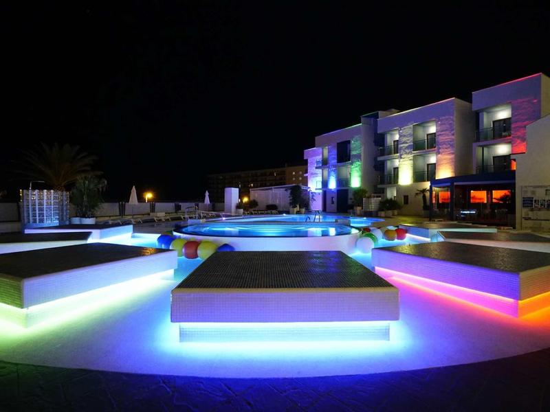 Moderner Pool mit leuchtenden Bänken und bunten LED-Lichtern, umgeben von Gebäuden bei Nacht.
