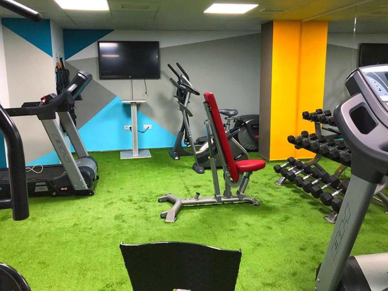 Modernes Fitnessstudio mit grünen Teppichboden, Trainingsgeräten und Hanteln an der Wand.