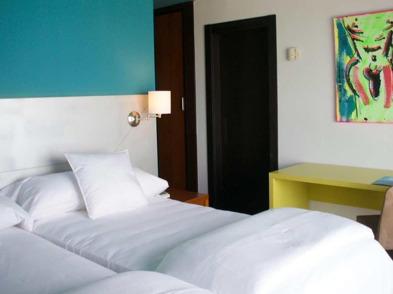 Modernes Schlafzimmer mit zwei Einzelbetten, weißen Bettwaren, grünem Tisch und farbenfrohem Bild.