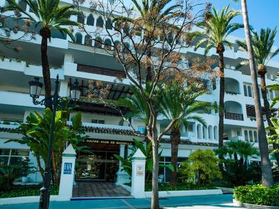 Mehrstöckiges weißes Gebäude mit Balkonen, Palmen und grünen Pflanzen davor bei blauem Himmel.