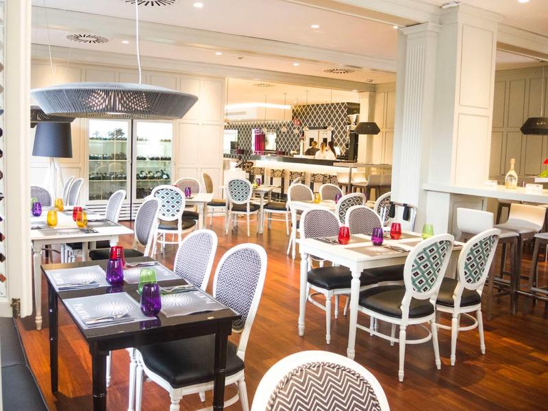 Helles Restaurant mit weißen Stühlen, Holzboden, gedeckten Tischen und moderner Bar im Hintergrund.