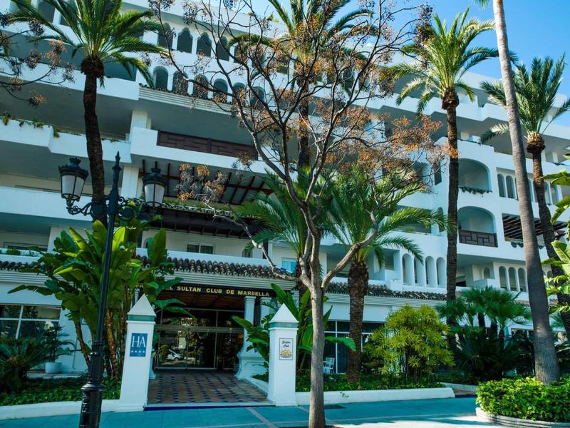 Mehrstöckiges weißes Gebäude mit Balkonen, Palmen und grünen Pflanzen davor bei blauem Himmel.