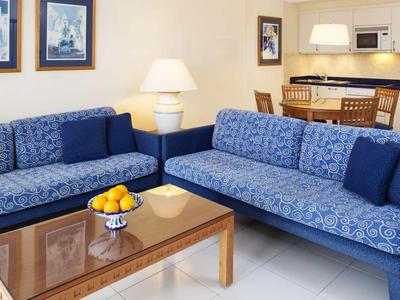 Modern eingerichtetes Wohnzimmer mit blauen Sofas, Holztisch und angrenzender Küchenzeile.