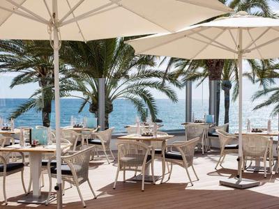 Außenbereich eines Cafés mit weißen Sonnenschirmen, Stühlen und Tischen am Meer mit Palmen