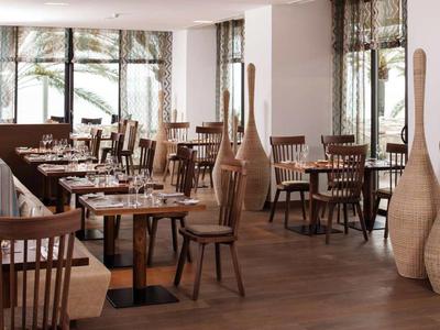 Gemütlicher, hell beleuchteter Speiseraum mit Holzstühlen und Tischen, große Vasen neben Fenstern.