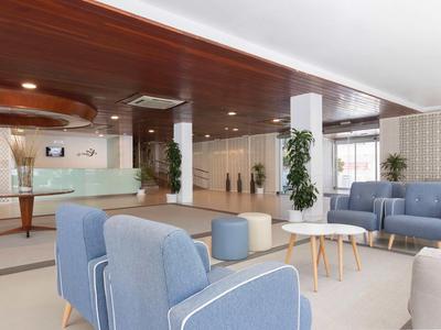 Světlá hala s moderními modrými křesly, pohovkami, dřevěnými stolky a rostlinami.