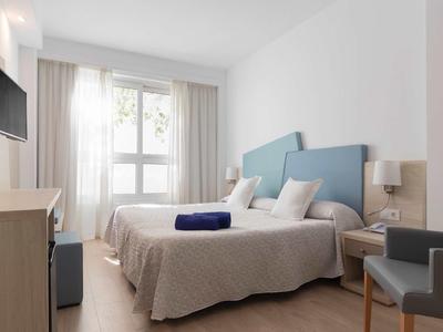 Světý hotelový pokoj s manželskou postelí, televizí, skříní a posezením u okna.