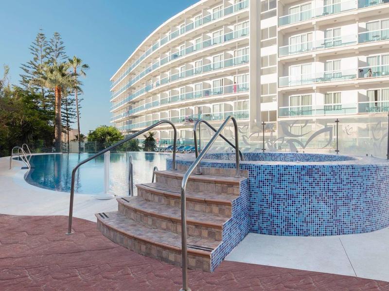 Moderner Außenpool mit runden Treppen und blauen Mosaikfliesen vor einem gebogenen Hotelgebäude.