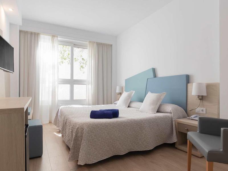 Helles Schlafzimmer mit zwei Einzelbetten, blauen Kopfteilen, grauen Decken und Fenster mit Vorhängen.