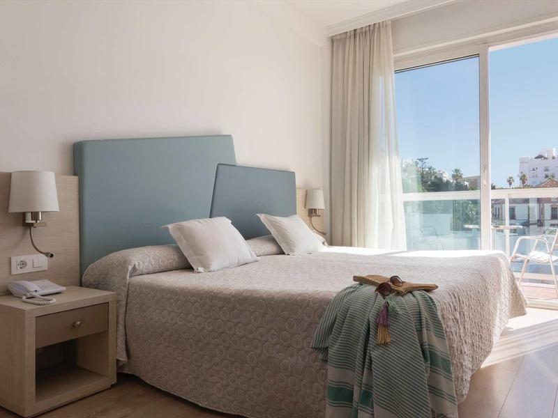 Helles Schlafzimmer mit Doppelbett, blauem Kopfteil, Nachttisch und großem Fenster mit Blick nach draußen.