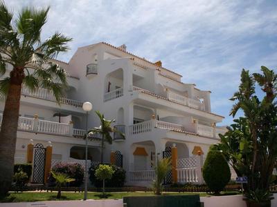 Immeuble d'hôtel blanc à plusieurs étages avec balcons, entouré de palmiers et ciel bleu.
