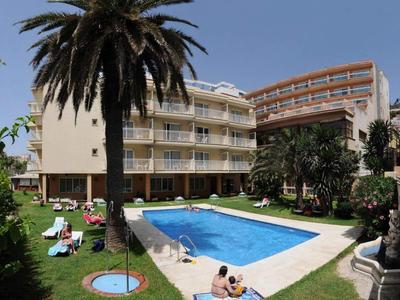 Hotelgebäude mit Pool, Palmen und Liegestühlen in einem grünen Garten unter klarem Himmel.