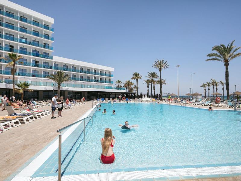 Großer Pool mit blauer Wasserfläche, umgeben von Sonnenliegen, Palmen und einem mehrstöckigen Hotel.