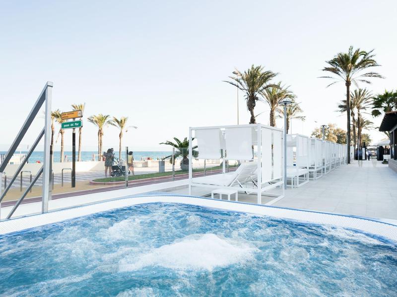 Whirlpool mit blubberndem Wasser, weißen Fliesen und Palmen am sonnigen Strand im Hintergrund.