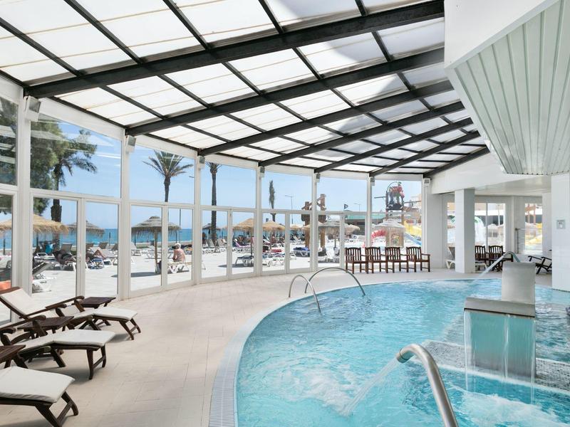 Großer Innenpool mit Glasdach, Liegestühlen und Blick auf Palmen und Meer dahinter.
