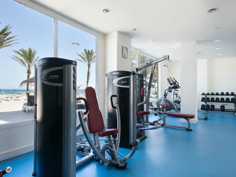 Helles Fitnessstudio mit blauer Matte, Kraftgeräten, Hanteln und Meerblick durch große Fenster.