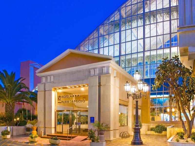 Eingang eines modernen Gebäudes mit Glasfassade, beleuchtet bei Abenddämmerung, umgeben von Pflanzen.