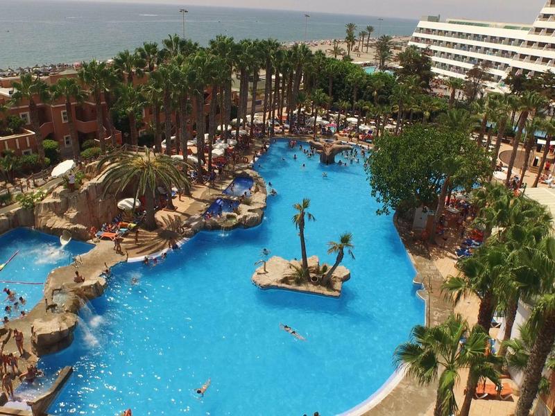 Großer Hotelpool mit Palmen, Sonnenschirmen und angrenzendem Strand am Meer im Hintergrund.