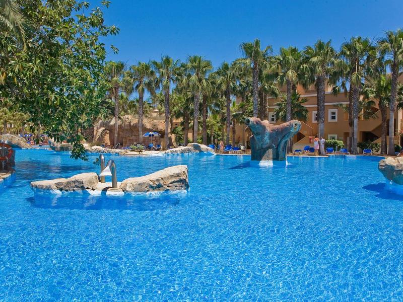 Großer blauer Pool mit Wasserspielen, umgeben von hohen Palmen und braunen Hotelgebäuden.