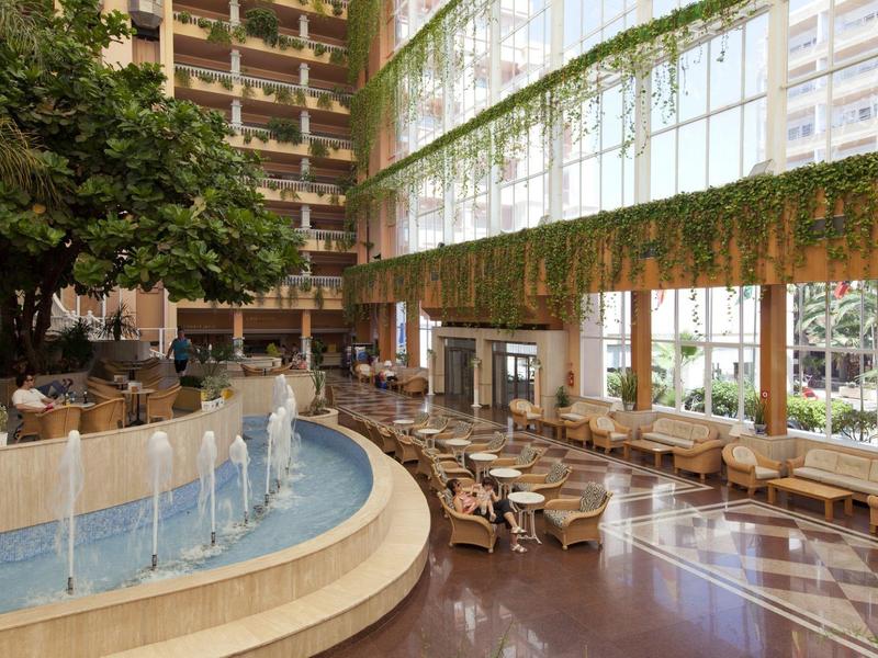 Großer Innenraum mit Wasserbrunnen, vielen Sitzplätzen und viel Grün an hohen Fensterfronten.