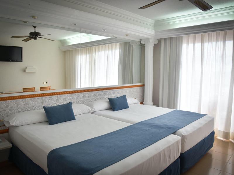 Helles Schlafzimmer mit zwei Einzelbetten, blauen Akzenten und großen, lichtdurchfluteten Fenstern.