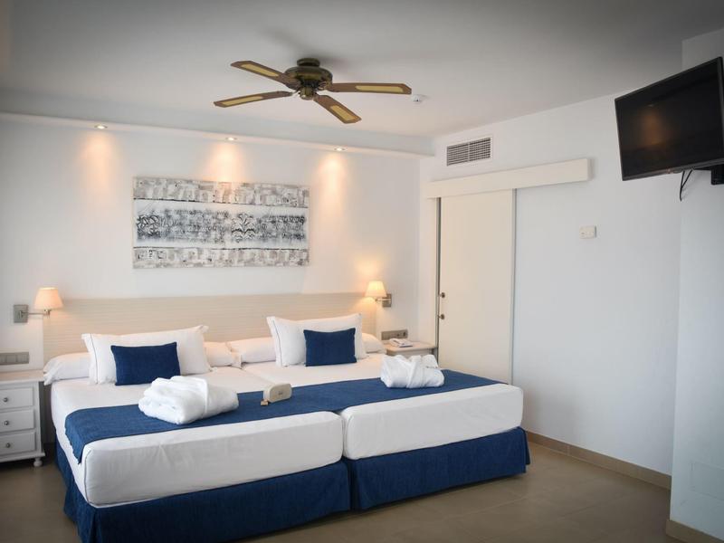 Helles Hotelzimmer mit zwei großen Betten, Ventilator, Wandbild und an der Wand montiertem Fernseher.