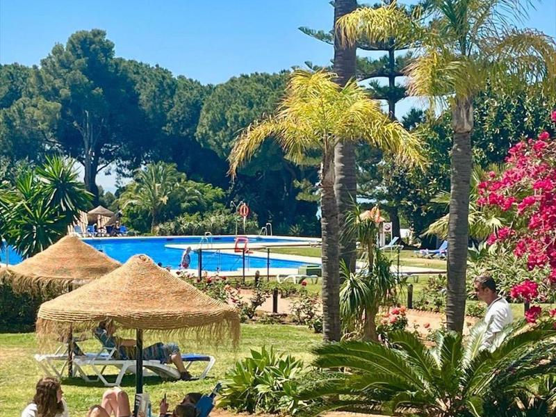 Grüne Gartenanlage mit Palmen, Sonnenschirmen, Liegestühlen und blauem Pool im Hintergrund.