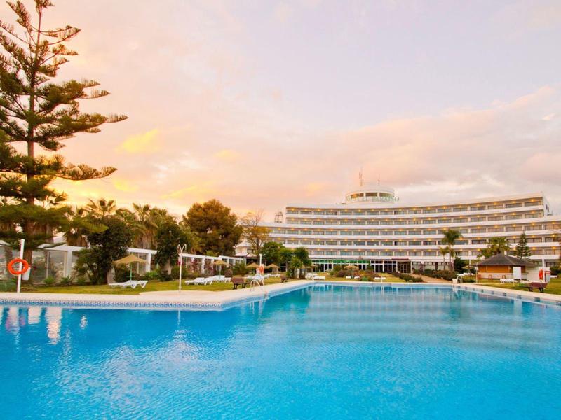 Großer blauer Pool vor weißem, mehrstöckigem Hotel mit Palmen und Sonnenschirmen bei Sonnenuntergang.