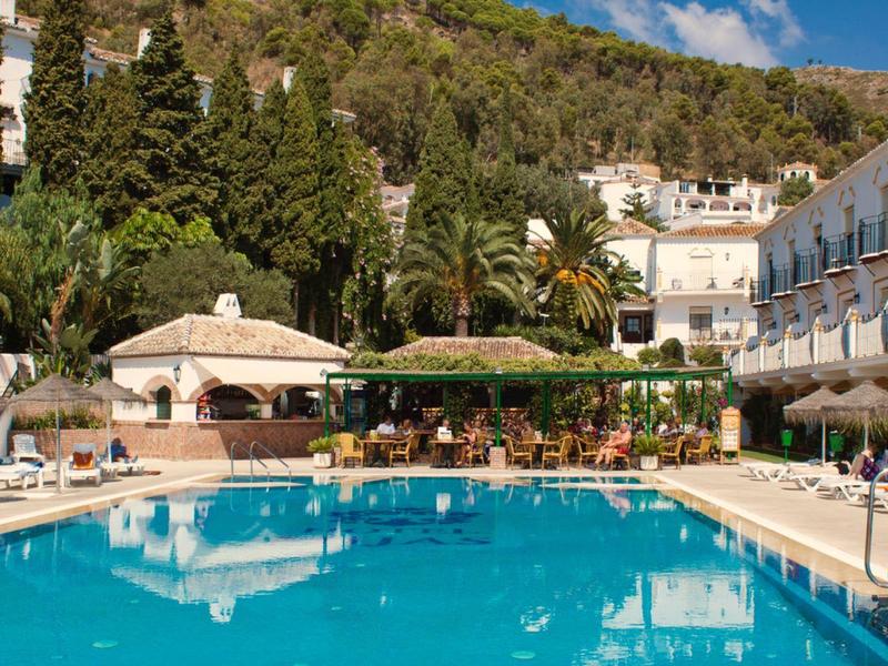 Großer blauer Hotelpool mit Liegestühlen, Sonnenschirmen und umgeben von grünen Bäumen und Gebäuden.