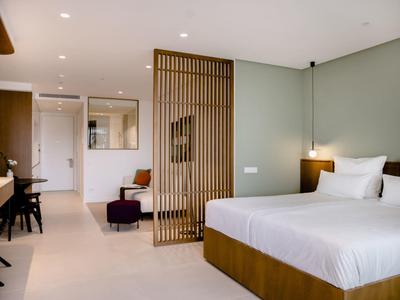 Modernes Hotelzimmer mit großem Bett, Holzmöbeln, Raumteiler und dezentem grünen Wandton.