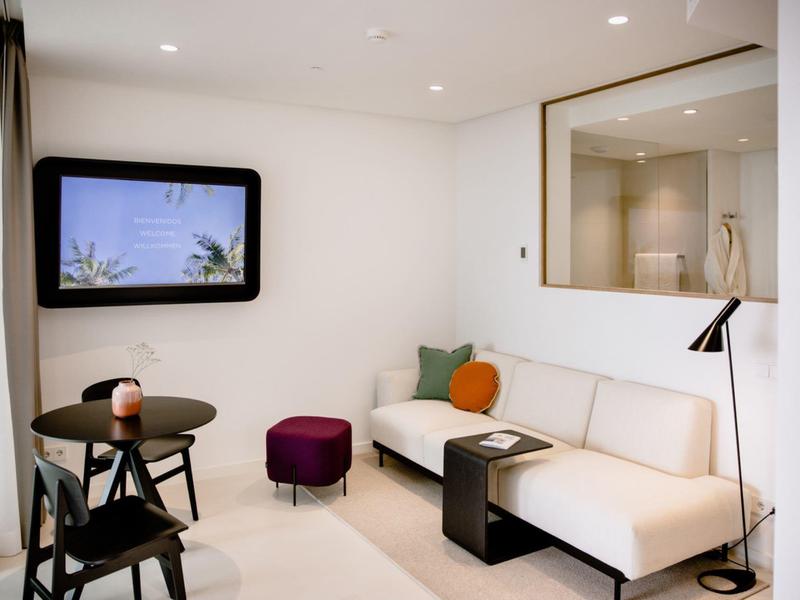 Modernes Wohnzimmer mit weißem Sofa, bunten Kissen, schwarzem Beistelltisch und Fenster mit Blick auf Palmen.