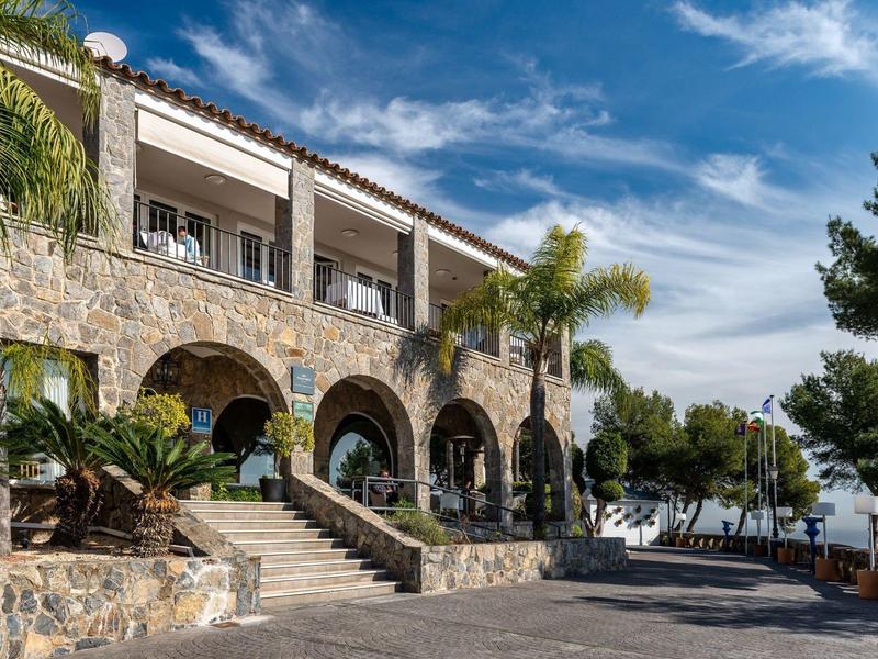 Stein Gebäude mit Bögen und Terrasse, umgeben von Palmen und Meerblick.