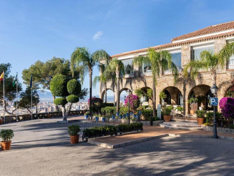 Hotel mit Arkadengängen und Palmen bei sonnigem Himmel neben einem Parkplatz