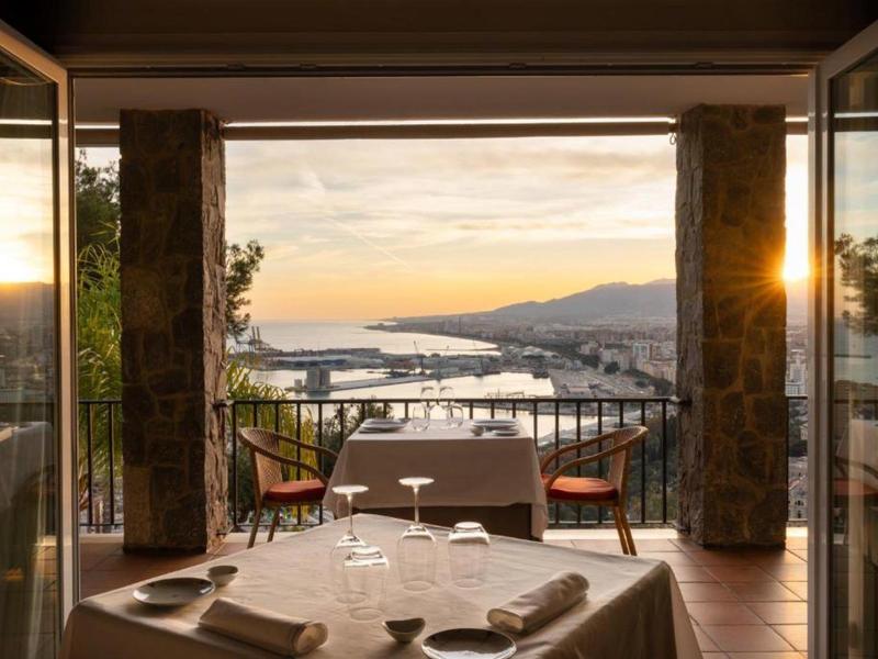 Terrasse mit Blick auf einen Hafen und Berge bei Sonnenuntergang, zwei Stühle und ein gedeckter Tisch.