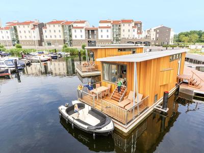 Hausboot mit Holzterrasse und kleinem Motorboot auf ruhigem Wasser in einem Hafen mit Gebäuden.