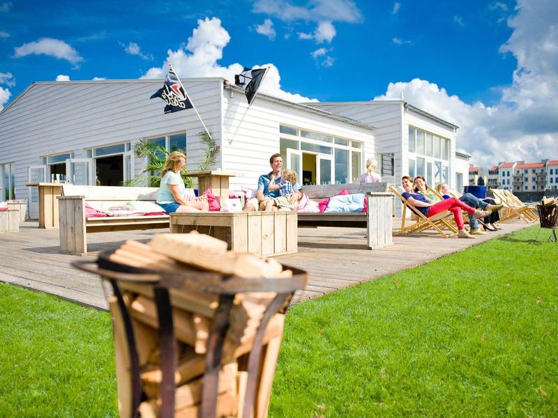 Modernes Haus mit großer Holzterrasse, Liegestühlen und Menschen bei sonnigem Wetter im Grünen.
