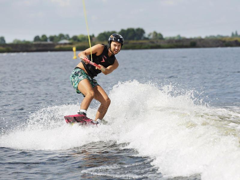 Mann macht Wakeboard-Fahren auf See, trägt Helm und Shorts, Wasser spritzt hinter ihm auf.