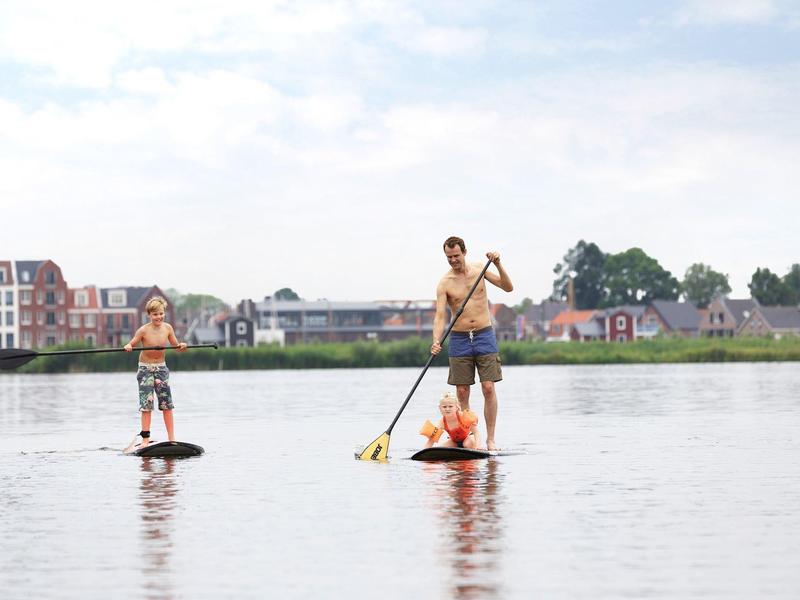 Zwei Personen auf Stand-Up-Paddle-Boards auf ruhigem Wasser vor Ufer mit Häusern und Bäumen.