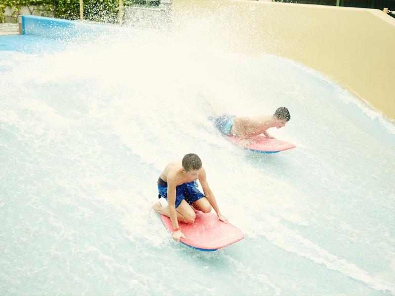 Zwei Jungen surfen auf bunten Boards in einem Wasserpark mit spritzenden Wellen.