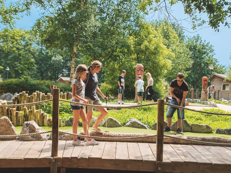 Kinder spielen Minigolf auf einem grünen Platz mit Holzsteg und Bäumen im Hintergrund.