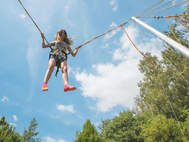 Ein Kind schaukelt hoch an Seilen, umgeben von grünem Laub und blauem Himmel mit Wolken.